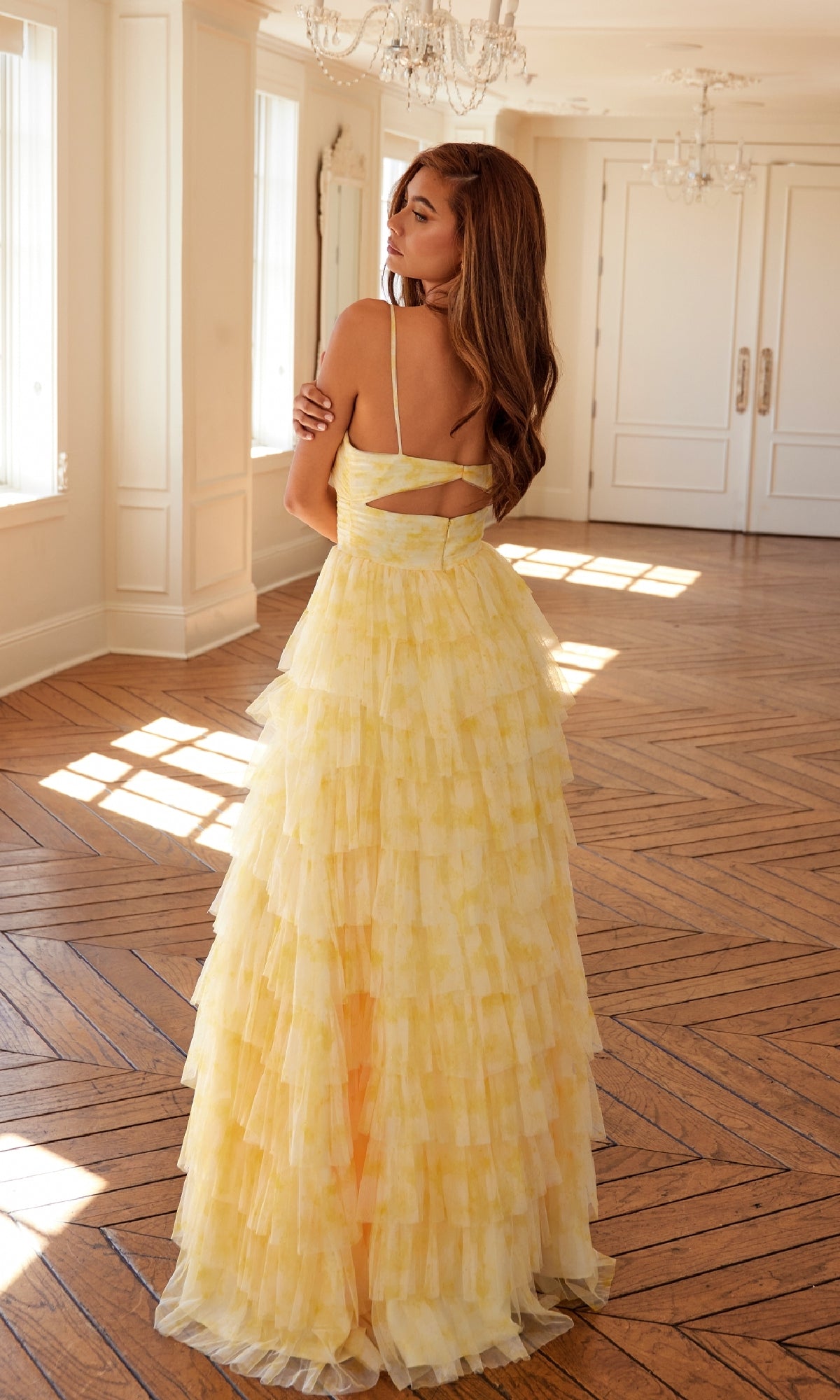 A woman stands indoors on herringbone wood flooring, facing away in the Juliet JT2508A Long Yellow Ruffled Prom Dress with a flowing A-line skirt, her head turned to the side.
